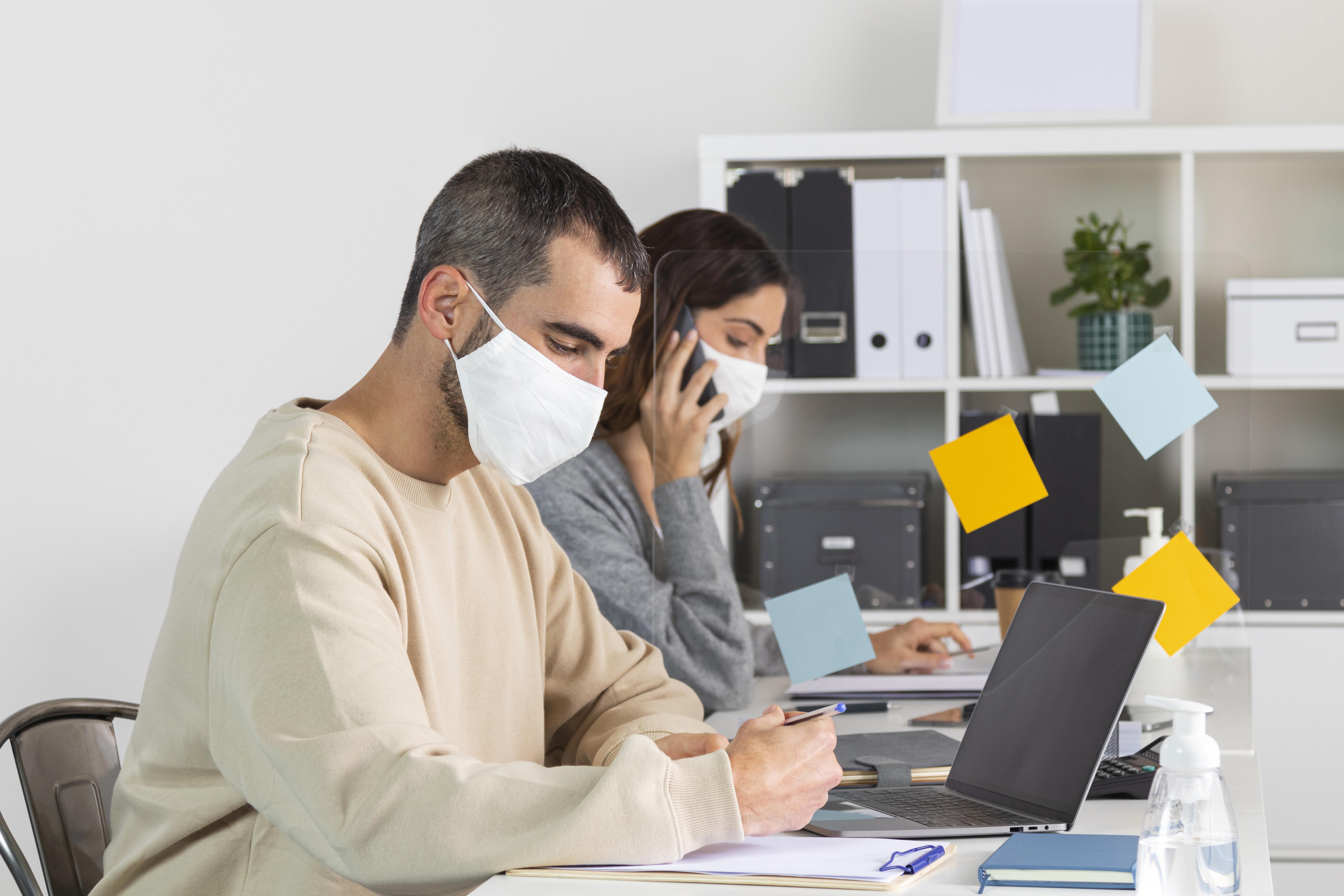 A-man-and-woman-wearing-masks-sit-at-a-desk-engaged-in-conversation-or-work-highlighting-safety-and-collaboration.